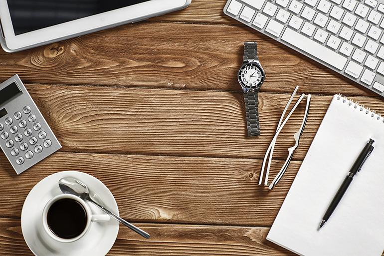 Photo of a coffee, a calculator, a pair of glasses, a watch, a notepad, a keyboard and tablet on a wooden desk.